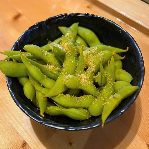 A black bowl filled with bright green edamame pods, sprinkled with white and yellow seasoning, on a wooden table.