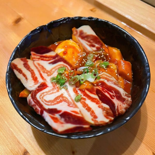 A black bowl of Korean tteokbokki with sliced pork belly, green onions, and sesame seeds.