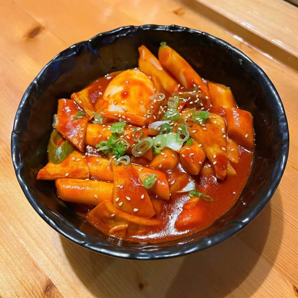 A black bowl of Izziban's Tteokbokki, Korean spicy rice cakes, topped with sesame seeds and scallions.