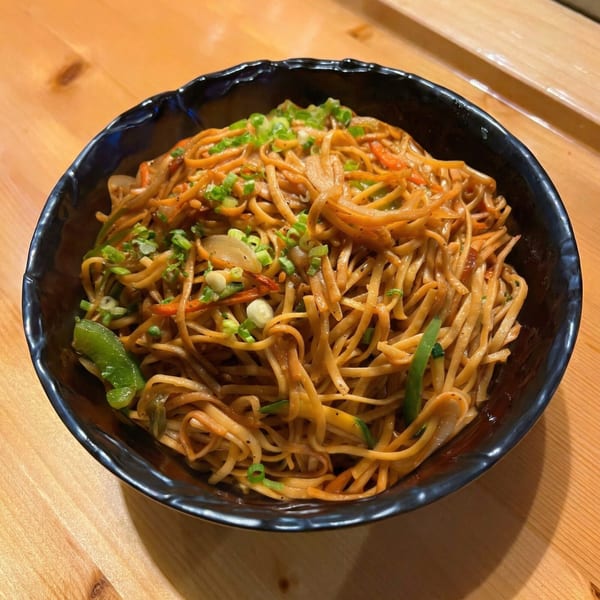 A black bowl of Korean BBQ noodles with vegetables and green onions on a wooden table.