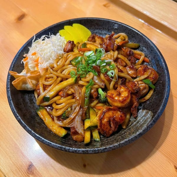 A black bowl of Korean BBQ udon noodles with shrimp, vegetables, and a side of pickled radish.