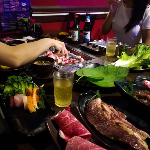 Diners cooking Korean BBQ at Izziban, with raw meats, sashimi, and drinks on the table.