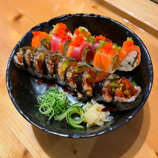 A black plate holds two rows of colorful sushi rolls, garnished with ginger, cucumber, and scallions.