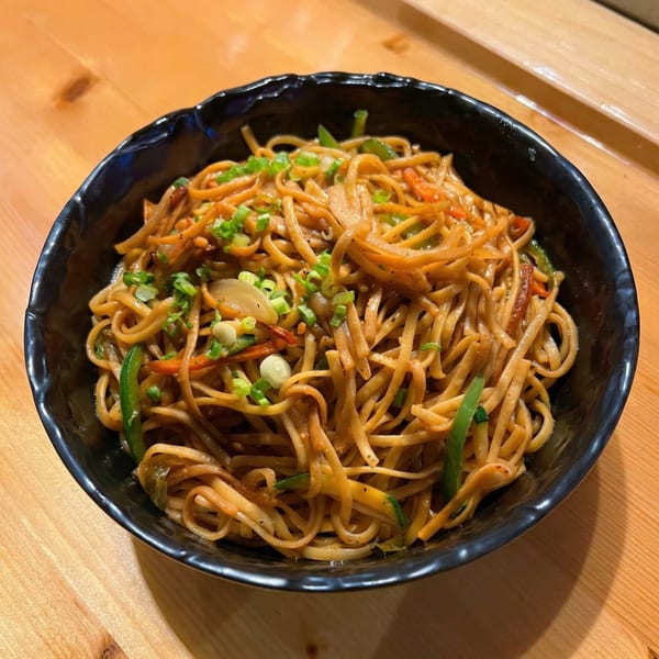 A black bowl of Korean BBQ noodles with vegetables and green onions on a wooden table.