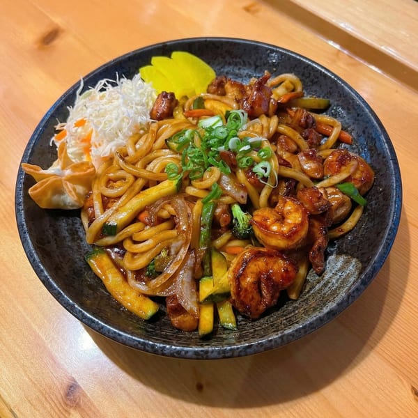 A black bowl of Korean BBQ udon noodles with shrimp, vegetables, and green onions.